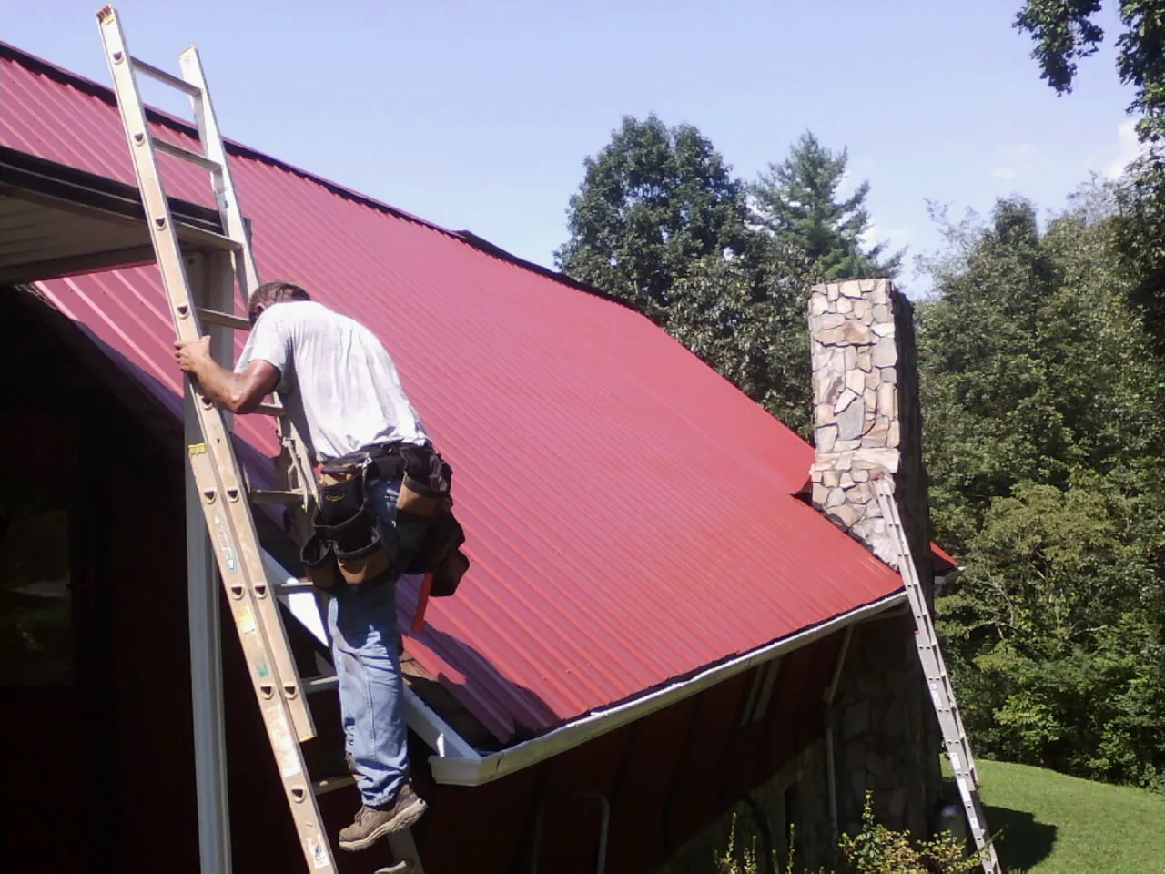 Roofer on a ladder beside a red metal roof and stone chimney — Soffit & Fascia Repair by Roofing Brothers in Sabattus