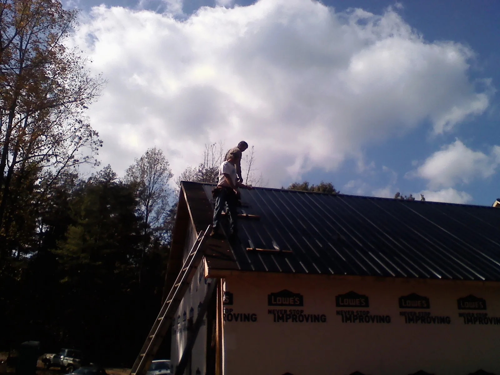 Roofer fastening dark metal panels high against a cloudy sky, from a Roof Inspection job in Sabattus